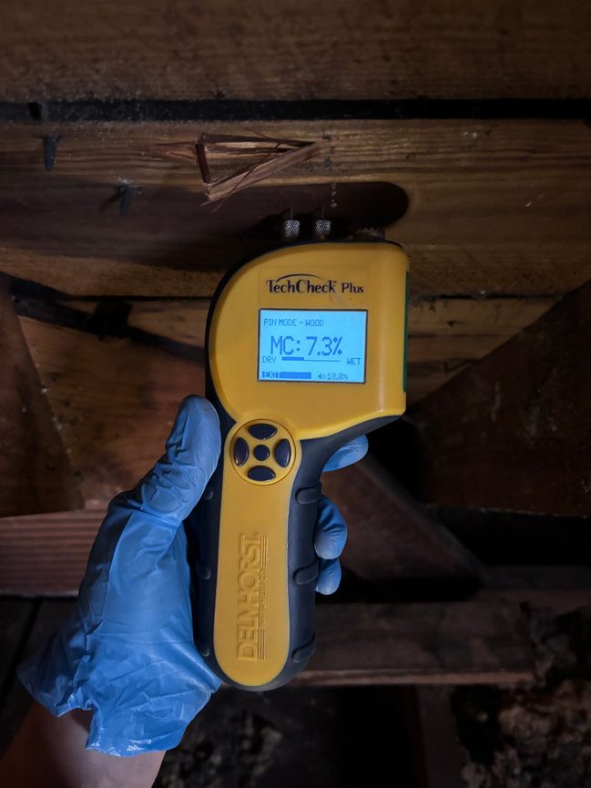A gloved hand holds a pin-type moisture meter against wood framing in a dark under-floor space, showing a 7.3 percent moisture content reading.