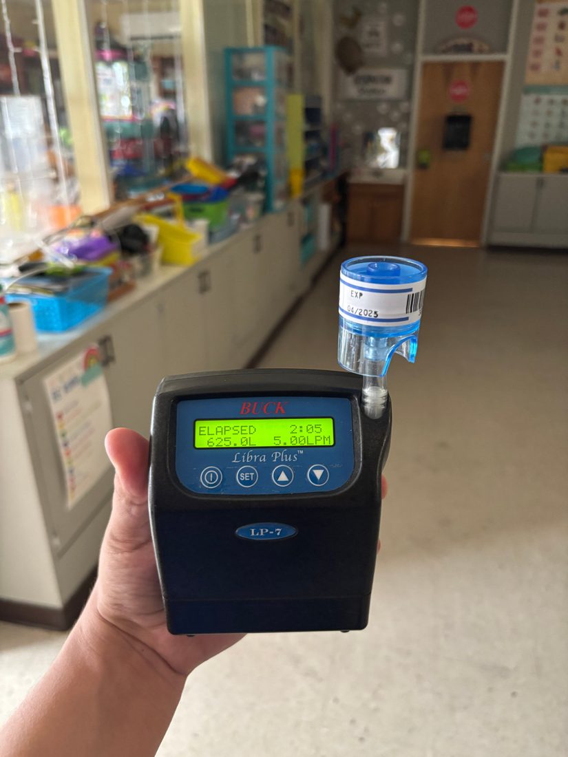 A hand holds a portable air sampling pump fitted with a spore trap cassette inside an occupied classroom.